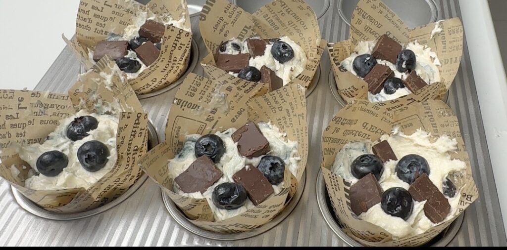 Six muffin liners filled with blueberry chocolate batter topped with chocolate chunks and blueberries before baking
