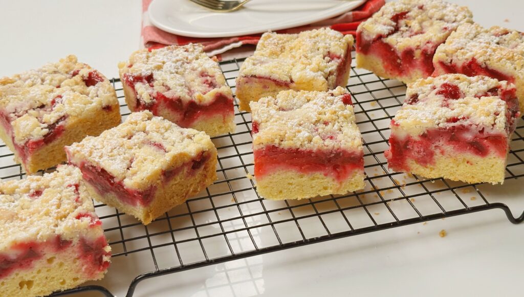Squares of plum streusel cake on a black cooling rack showing the soft base, red plum layer and crumb topping