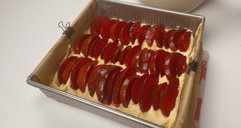 Sliced red plums arranged in overlapping rows over cake batter in a square baking tin before baking