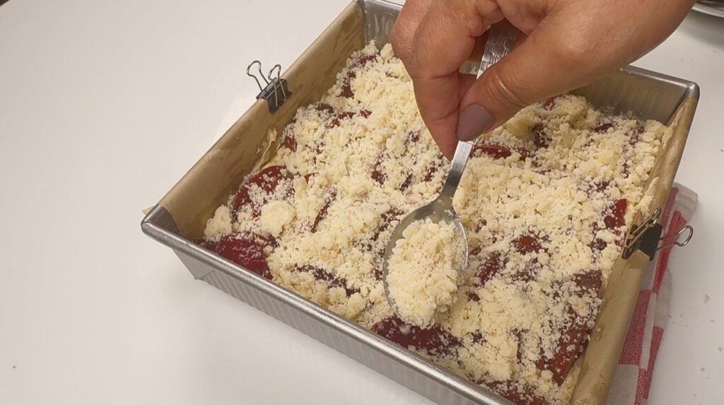 Buttery streusel crumbs being spooned over plums in a square baking tin before baking