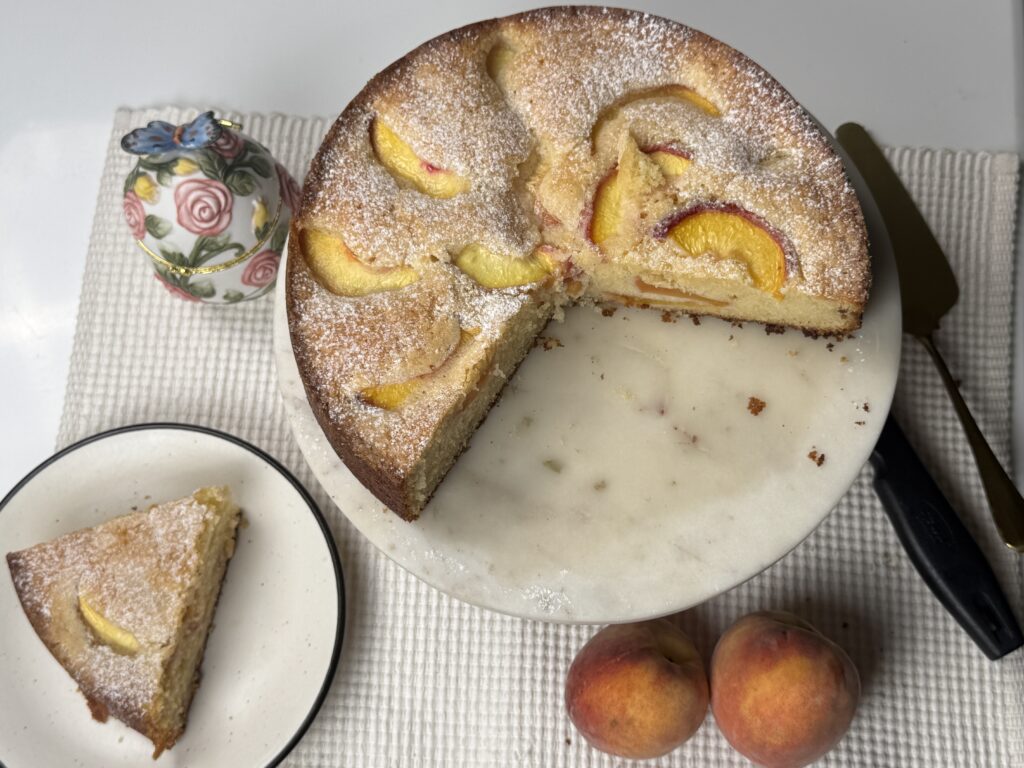 Overhead view of peach butter cake with a plated slice, knife, and fresh peaches nearby