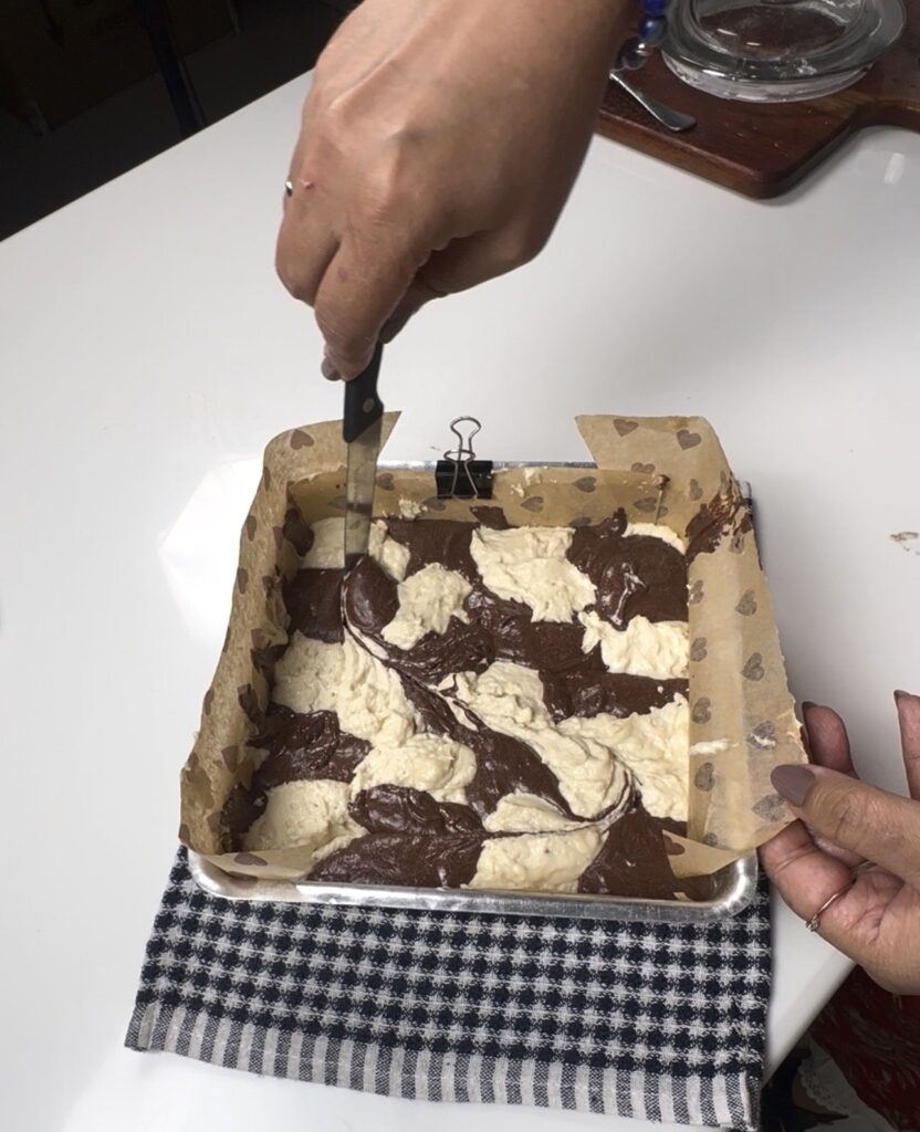 Swirling vanilla and chocolate batter with a knife in a square cake tin