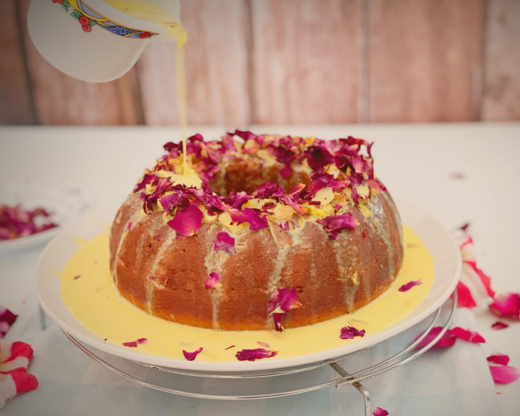 Pouring saffron rasmalai milk over an eggless bundt cake topped with rose petals