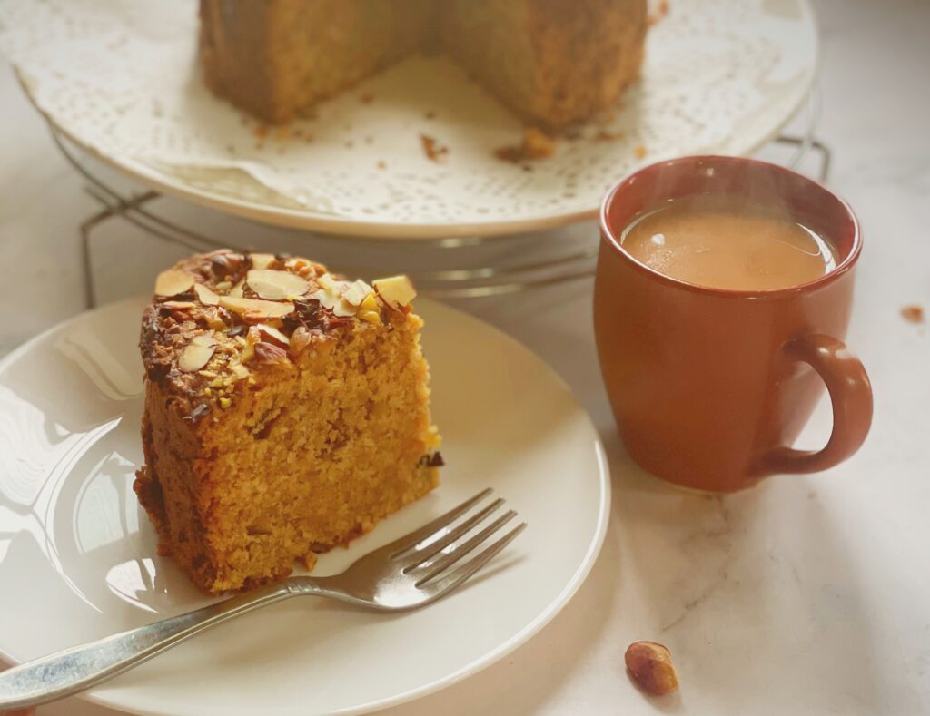 Slice of eggless mawa cake on a plate with a fork and a cup of chai