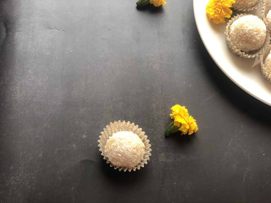 One coconut ladoo in a paper liner next to a marigold