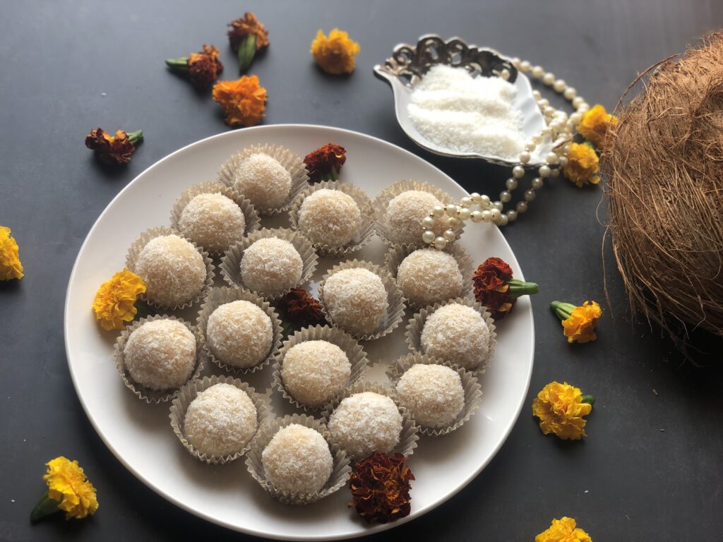 Coconut ladoos on a white plate with marigolds, a fresh coconut and a bowl of desiccated coconut