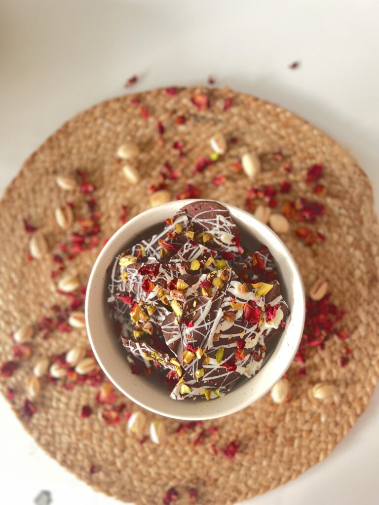 Bowl of rose pistachio chocolate bark on a jute mat surrounded by pistachios and rose petals