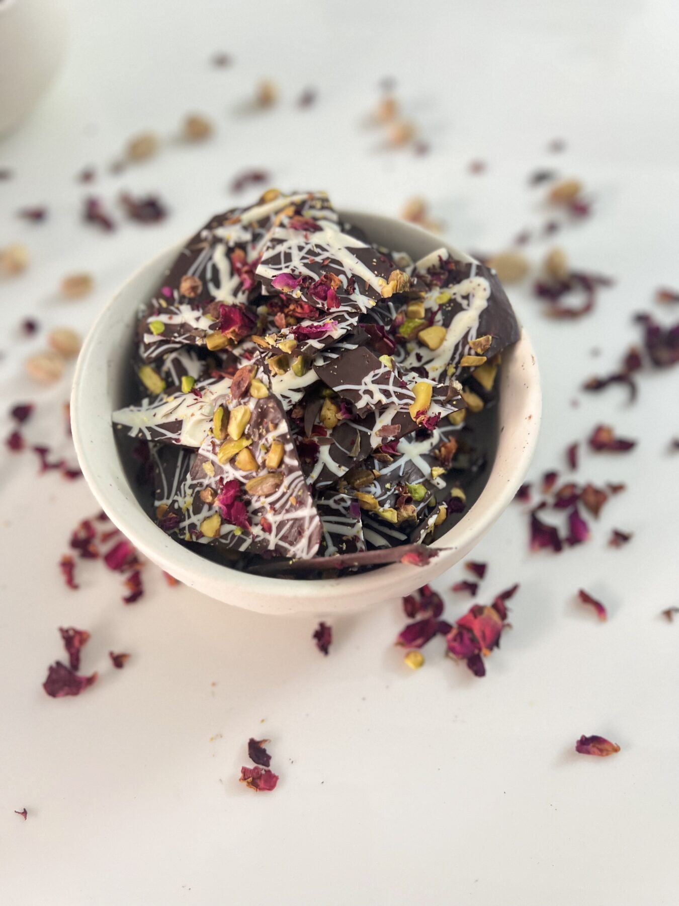 Pieces of dark chocolate bark with white chocolate drizzle, pistachios and rose petals in a white bowl