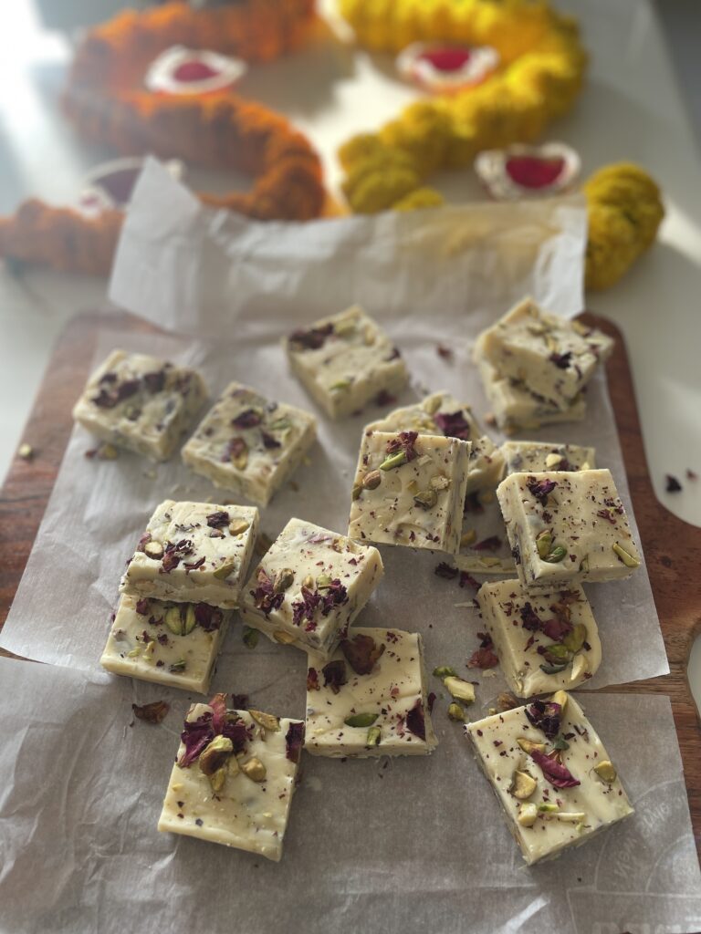 Squares of rasmalai fudge on butter paper with a marigold rangoli in the background