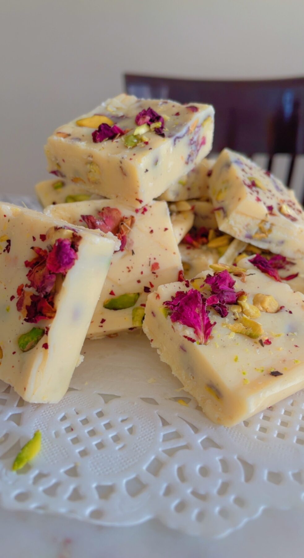 Stacked squares of rasmalai white chocolate fudge with rose petals and pistachios on a lace doily