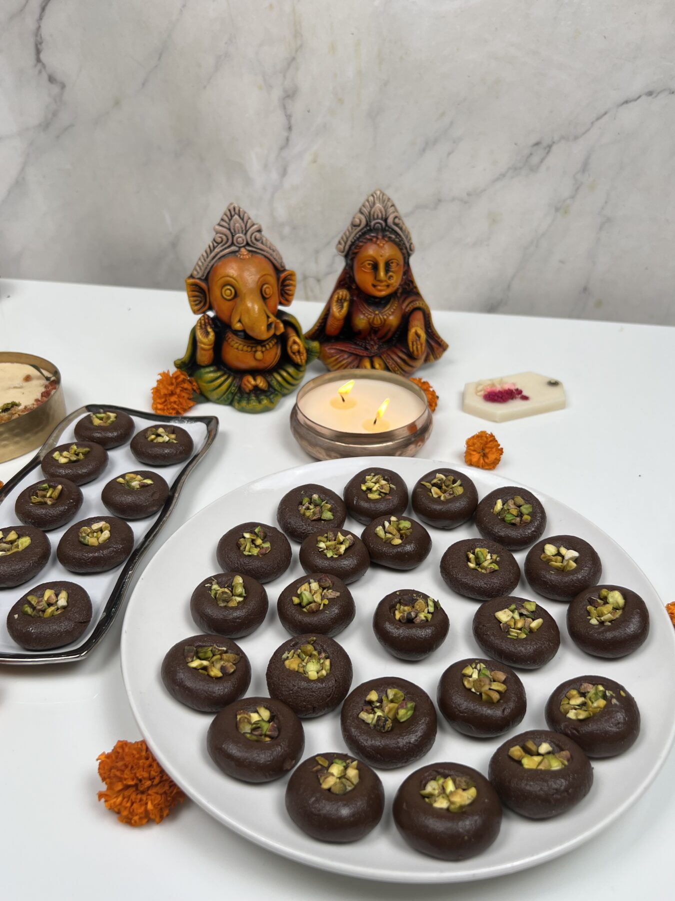 Chocolate pedas with pistachios on a plate for Diwali, with Ganesha and Lakshmi idols and a diya