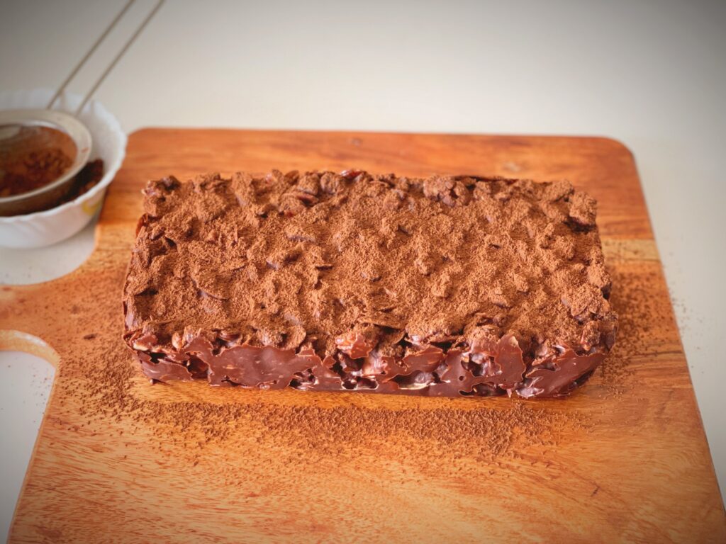 Whole no-bake chocolate biscuit cake dusted with cocoa powder on a wooden board